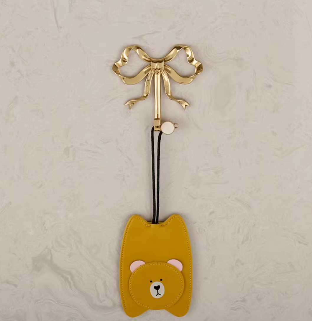 Golden bow tie brass wall hook holding a yellow bear-shaped charm on a marble background.